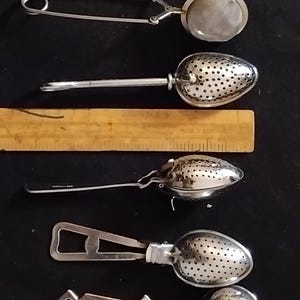 Peut inclure: Une collection de six infuseurs à thé en métal argenté de formes et styles variés, incluant des modèles en maille et perforés, disposés à côté d'une règle en bois mesurant environ 15 cm de longueur pour servir d'échelle de référence.