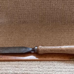 May include: A vintage wood-handled chisel with a metal blade, resting on a wooden ruler.