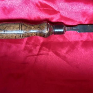 May include: A wooden handled chisel with a metal blade. The handle is brown with a dark wood grain and a metal cap on the end. The blade is silver and has a slight curve to it.