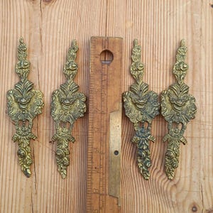 May include: Four ornate gold-toned metal decorative pieces with a floral and scroll design. The pieces are approximately 7.5 cm in length and are shown next to a ruler for scale.
