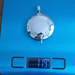 Könnte beinhalten: Ein glänzendes, silbernes, kreisförmiges Objekt mit einem kleinen, verstellbaren Stab und einer Feder. Es liegt auf einer blauen Waage, die ein Gewicht von 119 Gramm anzeigt.