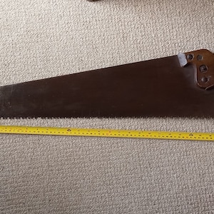 Può includere: Una sega a mano vintage con manico in legno e lama in metallo. La sega è lunga 68,58 cm e ha un metro a nastro giallo accanto.