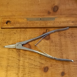 May include: A pair of silver metal pliers with long, pointed jaws and curved handles. A wooden ruler with measurement markings is in the background. The pliers are designed for precision work.