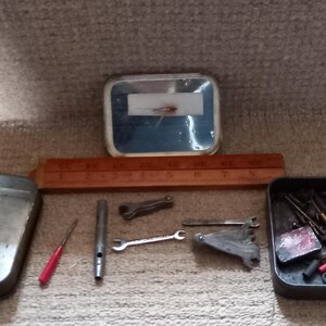Könnte beinhalten: Eine Sammlung von Vintage-Werkzeugen, darunter ein Lineal, Schraubenschlüssel und kleine Metallbehälter. Die Werkzeuge sind auf einer strukturierten Oberfläche angeordnet, was auf eine Werkstatt oder Reparaturumgebung hindeutet. Das Lineal ist in Zoll markiert.