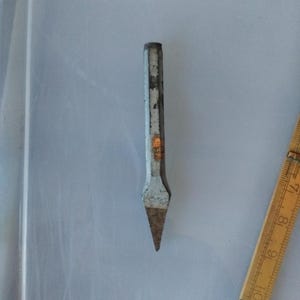 May include: A grey metal cold chisel with a worn finish and a pointed tip, positioned next to a yellow folding ruler showing measurements in centimeters, with a small orange label visible on the shaft of the tool.