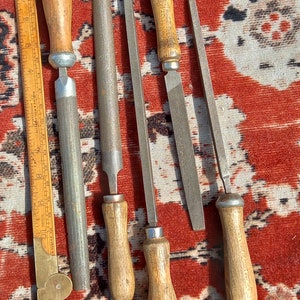 Może przedstawiać: Pięć metalowych pilników z drewnianymi uchwytami. Pilniki mają różne długości i różne grubości zębów. Pilniki leżą na powierzchni z czerwono-białym wzorem.