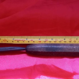 May include: A wooden-handled carving tool with a metal blade. The tool is about 7 inches long and is lying on a pink surface. A yellow measuring tape is visible in the background.