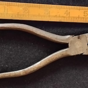 Op de afbeelding: Een tang van verweerd metaal met een textuurrijke grijze afwerking, geplaatst naast een gele houten vouwliniaal die laat zien dat het gereedschap een totale lengte heeft van ongeveer 14 cm, van het uiteinde van de handgreep tot de bek.