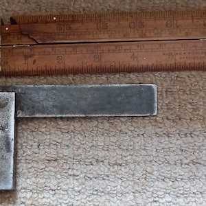 Op de afbeelding: Een metalen winkelhaak met een afgeschuinde rand en een gemarkeerde hoek. De winkelhaak ligt op een bruin oppervlak.