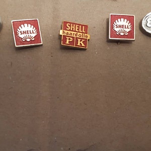 May include: Five vintage metal pins with logos. The pins are red, gold, and silver. The logos include "OK", "SHELL", "SHELL haardolie PK", "SHELL", and "ESSO".