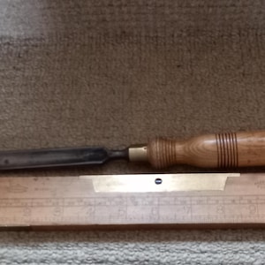 May include: A wooden and metal chisel with a brass ferrule and a wooden handle. The chisel is resting on a wooden ruler with a brass strip and markings.