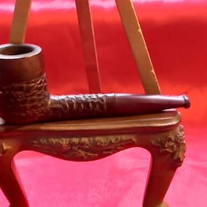 May include: A brown wooden pipe with a textured surface and a red mouthpiece. The pipe is resting on a decorative wooden stand.