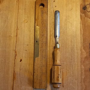 Op de afbeelding: Een houten beitel met een metalen blad. De beitel is gemarkeerd met "INSTRUCTOR" en is ongeveer 18 cm lang. De beitel ligt op een houten oppervlak naast een houten liniaal.