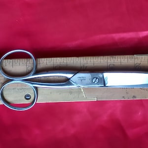 Może przedstawiać: Srebrne nożyczki z drewnianym linijką z tyłu. Linijka jest oznaczona w calach. Nożyczki mają małą śrubę w środku, a na ostrzu nadrukowane są słowa "America" i "Origin".