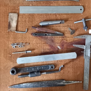 May include: A collection of various tools and metal objects, including a ruler, a file, a wrench, a screwdriver, a pair of pliers, a metal pin, and a metal rod. The tools are arranged on a wooden surface.