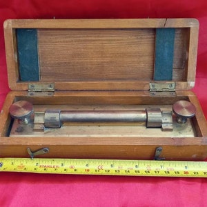 Puede incluir: Una herramienta de medición de latón con una escala graduada, en una caja de madera con tapa con bisagras. La herramienta mide aproximadamente 25 cm de largo.