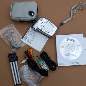 Pode incluir: Uma câmera de vídeo digital Vivitar DVR-510 prateada com uma capa de transporte preta, um tripé e um CD com instruções. A câmera tem uma tampa de lente e uma tela na parte de trás.