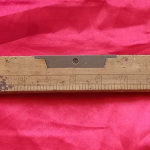 Puede incluir: Un nivel de madera con un nivel de burbuja de metal y una regla marcada en pulgadas del 1 al 9. El nivel está desgastado y tiene una pátina marrón.