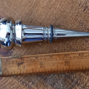 Puede incluir: Un tapón de botella de plata con una parte superior redondeada y una punta larga y puntiaguda. El tapón mide aproximadamente 7,5 cm de largo.