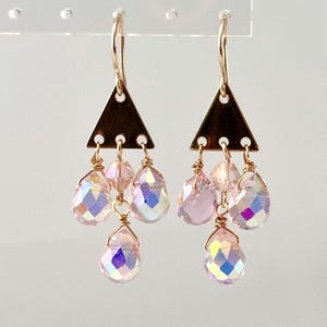 Puede incluir: Pendientes colgantes dorados con una parte superior triangular y tres cristales rosas iridiscentes en forma de lágrima colgando. Los pendientes tienen un diseño delicado, perfecto para añadir un toque de brillo a cualquier atuendo.