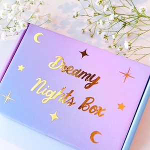 Puede incluir: Una caja de color lila y azul pastel con estrellas doradas, lunas y las palabras "Dreamy Nights Box". La caja está decorada con flores blancas.