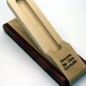 May include: A wooden pen holder with a sliding top. The holder is made of two types of wood, one light and one dark. The top of the holder is engraved with the text "Detective Sgt. Chris Caccibuillo".