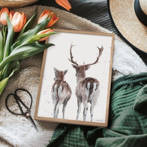 May include: A framed watercolour painting of two deer, one adult and one fawn, standing side by side. The deer are facing away from the viewer, with their tails up. The background is a light purple with white dots.