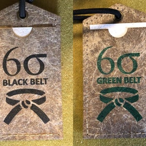 Könnte beinhalten: Zwei braune Lederetiketten mit einem weißen Papiereinsatz. Die Etiketten sind mit "66" nummeriert und haben den Text "Black Belt" und "Green Belt" aufgedruckt. Jedes Etikett hat einen schwarzen und grünen Gürtel aufgedruckt.