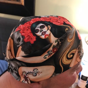 May include: A black and red bandana with a skull and floral design. The bandana features a woman with a sugar skull makeup design.