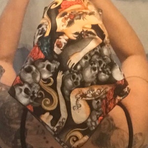 May include: A black fabric head wrap with a skull and crossbones pattern. The fabric features a white and gold design with a red rose and a woman's arm.