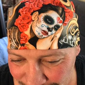 May include: A black bandana with a colorful design featuring a woman with a sugar skull face, red roses, and gold swirls.