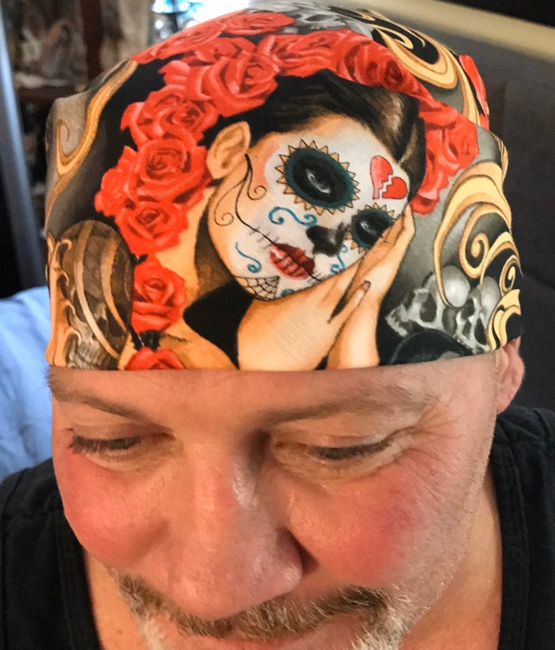 May include: A black bandana with a colorful design featuring a woman with a sugar skull face, red roses, and gold swirls.