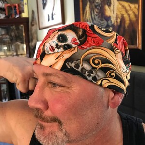 May include: A black and gold patterned bandana with a colorful sugar skull design. The bandana features a skull with a red heart, red roses, and intricate details.