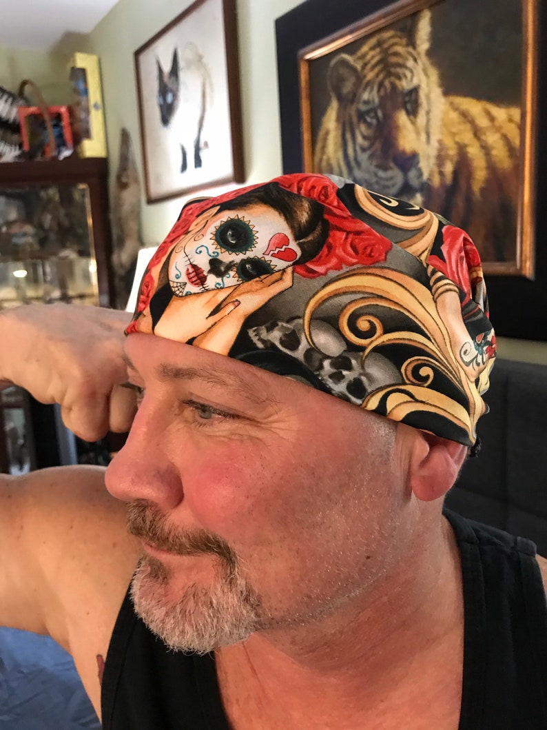 May include: A black and gold patterned bandana with a colorful sugar skull design. The bandana features a skull with a red heart, red roses, and intricate details.