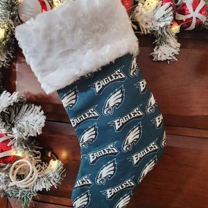 May include: A green and white Christmas stocking with a white faux fur cuff. The stocking is decorated with a repeating pattern of the Philadelphia Eagles logo and the word "Eagles".