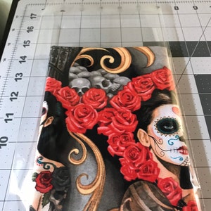 May include: A black and gold fabric with a design of two women with sugar skulls and red roses. The fabric is perfect for crafting projects like pillows, bags, and clothing.
