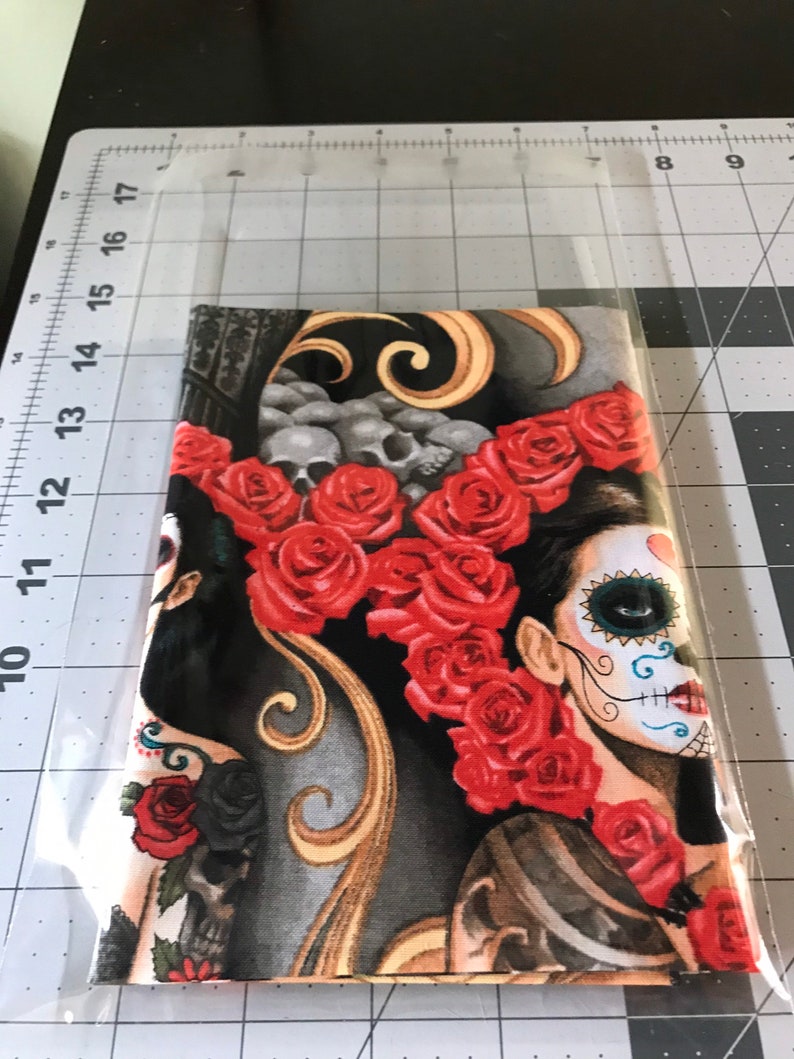 May include: A black and gold fabric with a design of two women with sugar skulls and red roses. The fabric is perfect for crafting projects like pillows, bags, and clothing.