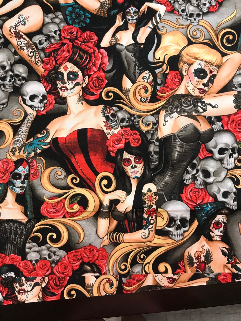 May include: A black fabric with a repeating pattern of sugar skulls, roses, and ornate swirls. The sugar skulls are decorated with colorful flowers and have black eyes. The roses are red and have green leaves. The swirls are gold and are arranged in a decorative pattern.