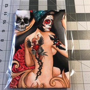 May include: A fabric panel with a colorful illustration of a woman with a skull face and tattoos. The woman is wearing a blue bandana and has a red heart tattoo on her back. The background is black with red roses.