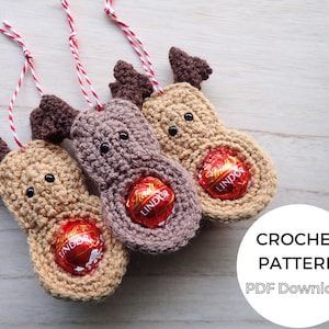 Puede incluir: Tres adornos de reno tejidos a crochet con hilo marrón y beige, cada uno con una trufa de chocolate Lindor. Los adornos tienen ojos negros y cuernos marrones. El fondo es una superficie de madera clara. CROCHET PATTERN PDF Download está escrito en texto blanco sobre un círculo negro.
