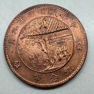 May include: A copper-colored coin with Chinese characters and a detailed relief of a building with figures. The coin's edge has a textured pattern, and the overall design suggests a historical or commemorative piece.