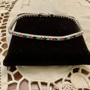 Peut inclure: Un bracelet en argent orné d'une rangée de pierres précieuses alternées rouges, vertes et bleues. Le bracelet est présenté sur un support en velours noir, mettant en valeur les couleurs vives des pierres. Le fermoir est visible.