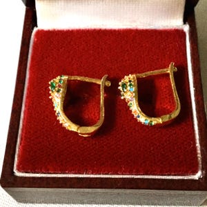 Peut inclure: Une paire de boucles d'oreilles dorées avec des pierres précieuses colorées. Les boucles d'oreilles sont présentées dans un écrin de bijoux doublé de velours rouge avec un cadre en bois brun foncé. Les pierres précieuses comprennent du vert, du bleu et de l'orange.