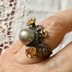 Peut inclure: Une bague en argent avec une grosse perle grise et des fleurs dorées avec des accents roses.