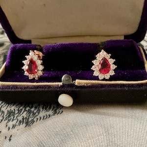 Peut inclure: Une paire de boucles d'oreilles avec une pierre rouge en forme de larme entourée d'un halo de petites pierres claires. Les boucles d'oreilles sont présentées dans un écrin en velours noir.