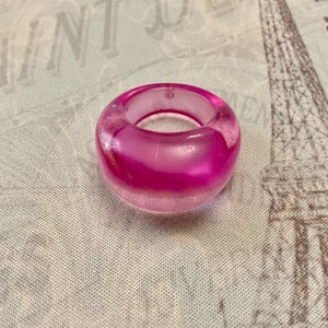 May include: A translucent, hot pink ring with a rounded shape. The ring appears to be made of resin or a similar material, with a smooth, glossy finish. The ring is a bold statement piece.
