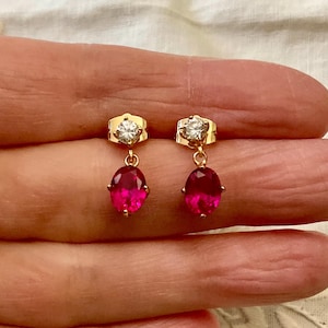 Peut inclure: Une paire de boucles d'oreilles pendantes dorées. Chaque boucle d'oreille est ornée d'une petite pierre précieuse ronde et transparente en haut, reliée à une pierre précieuse rose ovale vibrante. Les boucles d'oreilles sont présentées sur un fond neutre et doux.
