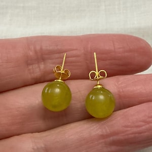 Peut inclure: Une paire de boucles d'oreilles à clous dorées, chacune ornée d'une pierre précieuse ronde vert clair. Les boucles d'oreilles sont présentées sur un fond neutre, soulignant leur design simple et leur couleur.