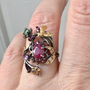 Peut inclure: Une bague en argent avec une grande pierre de rubis entourée de petites pierres roses. La bague présente un design délicat de branches avec des accents dorés et deux petits oiseaux.