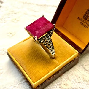 以下が含まれることがあります： 大きな長方形のルビー色の宝石が付いたシルバーリング。リングは装飾的な透かし細工のデザインで、ヴィンテージのジュエリーボックスに入っており、「AU ROY YVES ROUEN」の文字が書かれています。
